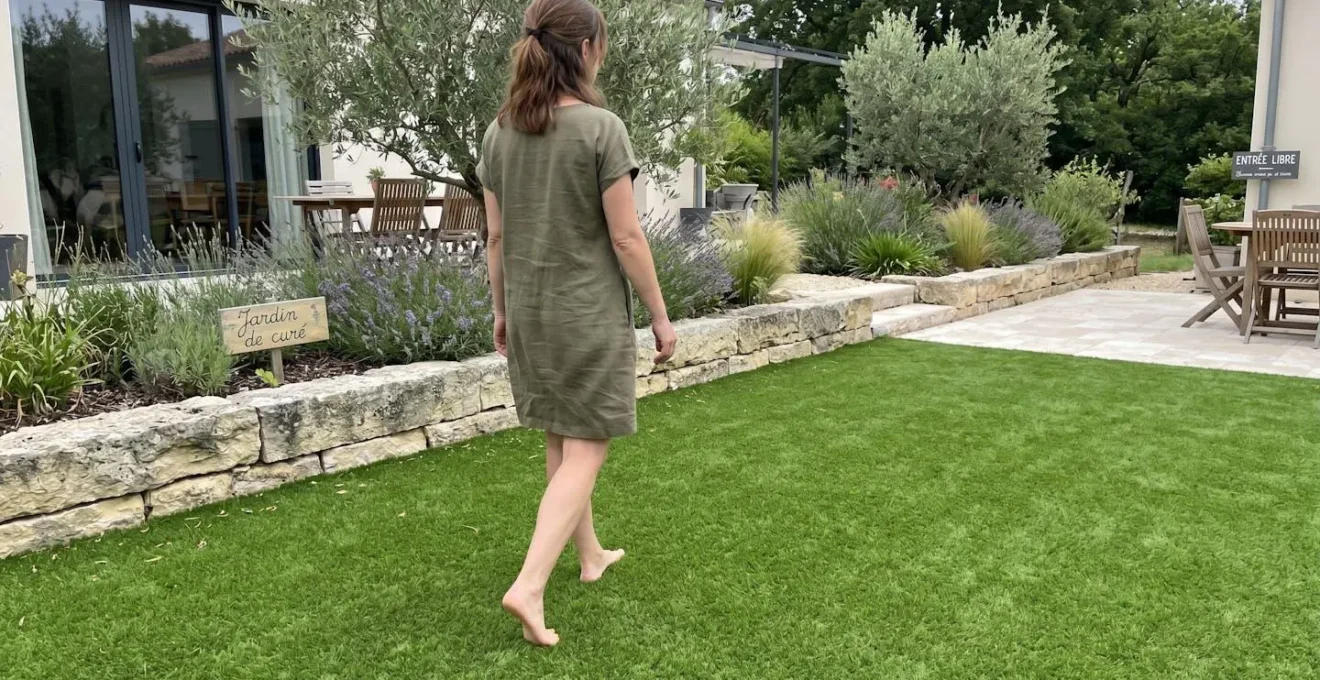 Personne vue de dos marchant pieds nus sur une pelouse synthétique, focus sur les jambes et les pieds, jardin résidentiel moderne en arrière-plan flouté, lumière naturelle douce